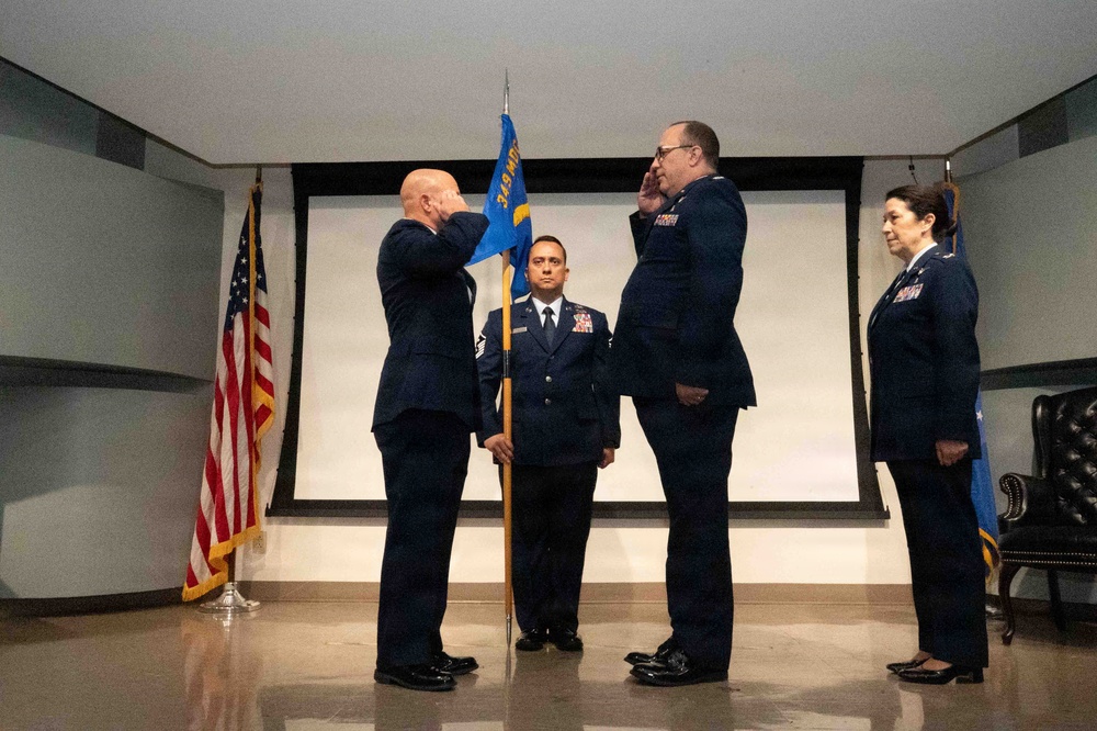 A formal U.S. Air Force military ceremony, likely a change of command or promotion, taking place indoors. Two senior officers are exchanging salutes while a third uniformed member holds a unit guidon flag in the center. A fourth officer stands at attention to the right near an Air Force flag.