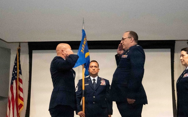 A formal U.S. Air Force military ceremony, likely a change of command or promotion, taking place indoors. Two senior officers are exchanging salutes while a third uniformed member holds a unit guidon flag in the center. A fourth officer stands at attention to the right near an Air Force flag.