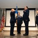 A formal U.S. Air Force military ceremony, likely a change of command or promotion, taking place indoors. Two senior officers are exchanging salutes while a third uniformed member holds a unit guidon flag in the center. A fourth officer stands at attention to the right near an Air Force flag.