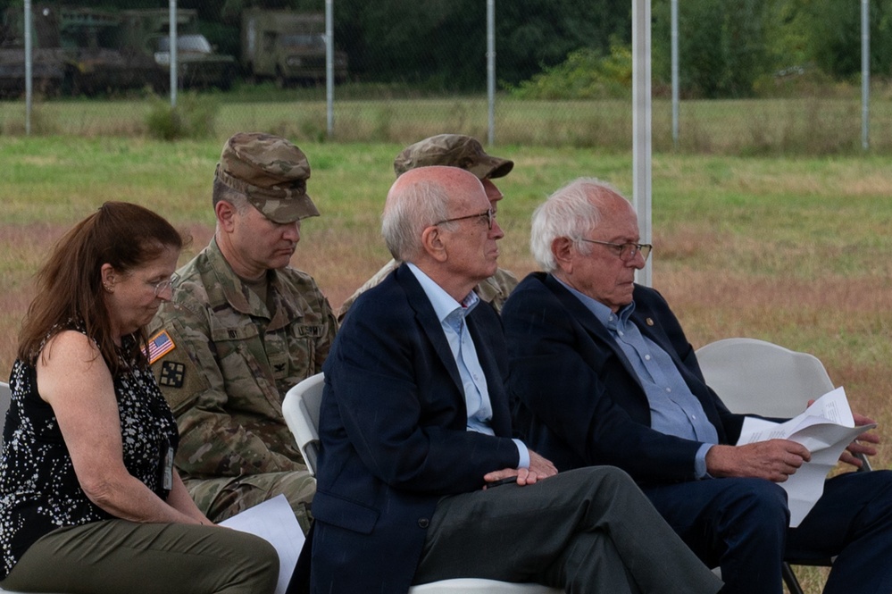 A group of individuals are seated in white folding chairs at what appears to be an outdoor military or official ceremony. The group includes two uniformed U.S. Army soldiers in camouflage (OCP) uniforms and three civilians, including a woman and two older men in business attire. Military vehicles and a fenced field are visible in the background.