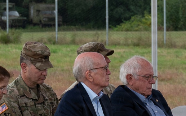 A group of individuals are seated in white folding chairs at what appears to be an outdoor military or official ceremony. The group includes two uniformed U.S. Army soldiers in camouflage (OCP) uniforms and three civilians, including a woman and two older men in business attire. Military vehicles and a fenced field are visible in the background.