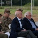 A group of individuals are seated in white folding chairs at what appears to be an outdoor military or official ceremony. The group includes two uniformed U.S. Army soldiers in camouflage (OCP) uniforms and three civilians, including a woman and two older men in business attire. Military vehicles and a fenced field are visible in the background.