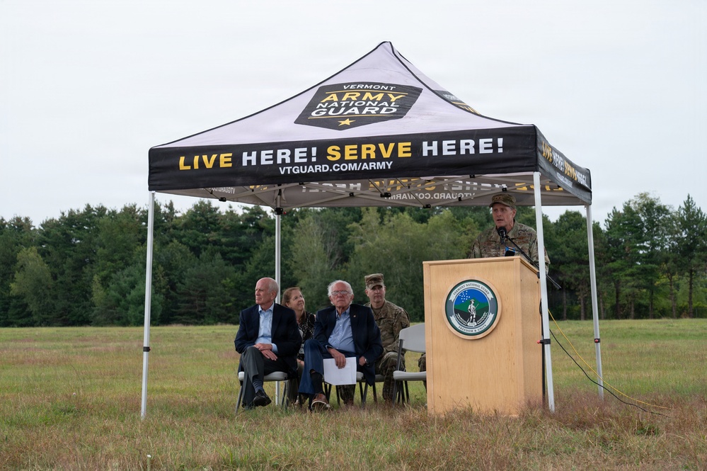 An outdoor ceremony is being held under a Vermont Army National Guard branded canopy tent in an open field. A uniformed military officer is speaking at a podium bearing what appears to be a Vermont National Guard seal, while several seated attendees — including civilians and at least one uniformed service member — listen. The setting appears to be a formal military or government event held in a rural, wooded area.
