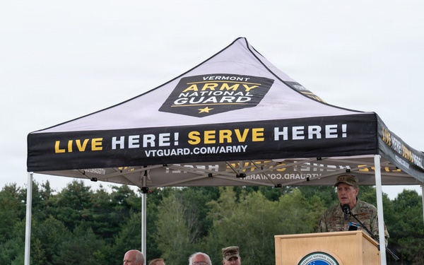 An outdoor ceremony is being held under a Vermont Army National Guard branded canopy tent in an open field. A uniformed military officer is speaking at a podium bearing what appears to be a Vermont National Guard seal, while several seated attendees — including civilians and at least one uniformed service member — listen. The setting appears to be a formal military or government event held in a rural, wooded area.