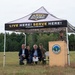 An outdoor ceremony is being held under a Vermont Army National Guard branded canopy tent in an open field. A uniformed military officer is speaking at a podium bearing what appears to be a Vermont National Guard seal, while several seated attendees — including civilians and at least one uniformed service member — listen. The setting appears to be a formal military or government event held in a rural, wooded area.