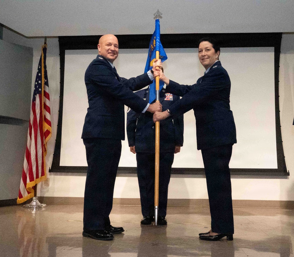 Two U.S. Air Force officers in dress blue uniforms are participating in a formal guidon/flag transfer ceremony, likely a change of command. A third uniformed service member stands at attention in the background. The ceremony takes place indoors on a stage with an American flag displayed to the left.