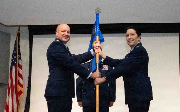 Two U.S. Air Force officers in dress blue uniforms are participating in a formal guidon/flag transfer ceremony, likely a change of command. A third uniformed service member stands at attention in the background. The ceremony takes place indoors on a stage with an American flag displayed to the left.