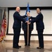 Two U.S. Air Force officers in dress blue uniforms are participating in a formal guidon/flag transfer ceremony, likely a change of command. A third uniformed service member stands at attention in the background. The ceremony takes place indoors on a stage with an American flag displayed to the left.
