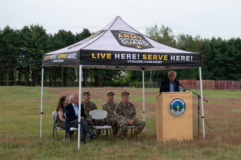 An outdoor event is taking place under a Vermont Army National Guard branded canopy tent in a grassy field. An elderly civilian man is speaking at a wooden podium bearing a Vermont National Guard seal, while five attendees — two civilians and three military personnel in camouflage uniforms — are seated in folding chairs beneath the tent. The setting appears to be a formal ceremony or announcement on National Guard grounds.