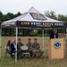 An outdoor event is taking place under a Vermont Army National Guard branded canopy tent in a grassy field. An elderly civilian man is speaking at a wooden podium bearing a Vermont National Guard seal, while five attendees — two civilians and three military personnel in camouflage uniforms — are seated in folding chairs beneath the tent. The setting appears to be a formal ceremony or announcement on National Guard grounds.