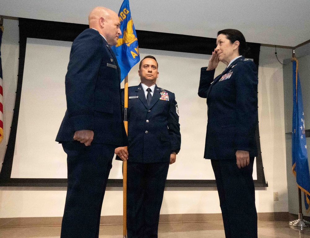 A formal U.S. Air Force military ceremony is taking place, likely a change of command or promotion ceremony. Three uniformed personnel are on a stage, with one officer on the right rendering a salute to a senior officer on the left, while a non-commissioned officer holds a unit guidon in the center. Military flags and a projection screen are visible in the background.