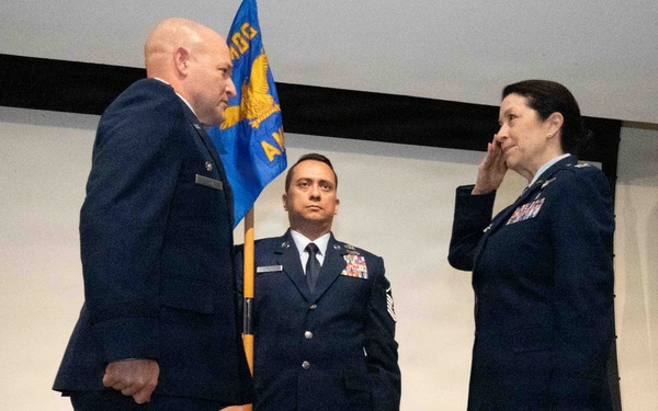 A formal U.S. Air Force military ceremony is taking place, likely a change of command or promotion ceremony. Three uniformed personnel are on a stage, with one officer on the right rendering a salute to a senior officer on the left, while a non-commissioned officer holds a unit guidon in the center. Military flags and a projection screen are visible in the background.