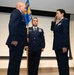 A formal U.S. Air Force military ceremony is taking place, likely a change of command or promotion ceremony. Three uniformed personnel are on a stage, with one officer on the right rendering a salute to a senior officer on the left, while a non-commissioned officer holds a unit guidon in the center. Military flags and a projection screen are visible in the background.