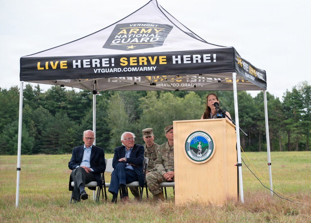 An outdoor ceremony is taking place under a Vermont Army National Guard branded canopy tent in a field. A woman is speaking at a podium bearing the Vermont National Guard seal, while four individuals — two in civilian attire and two in military camouflage uniforms — are seated behind her. The setting appears to be an official military or government event.