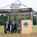An outdoor ceremony is taking place under a Vermont Army National Guard branded canopy tent in a field. A woman is speaking at a podium bearing the Vermont National Guard seal, while four individuals — two in civilian attire and two in military camouflage uniforms — are seated behind her. The setting appears to be an official military or government event.