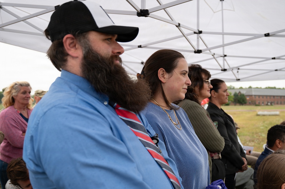 A group of people stand attentively under a white pop-up canopy tent at what appears to be an outdoor ceremony or event. The foreground features a bearded man in a blue shirt and striped tie wearing a black baseball cap, alongside a woman in a blue striped top with gold necklaces. A brick building and open field are visible in the background.