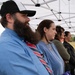 A group of people stand attentively under a white pop-up canopy tent at what appears to be an outdoor ceremony or event. The foreground features a bearded man in a blue shirt and striped tie wearing a black baseball cap, alongside a woman in a blue striped top with gold necklaces. A brick building and open field are visible in the background.