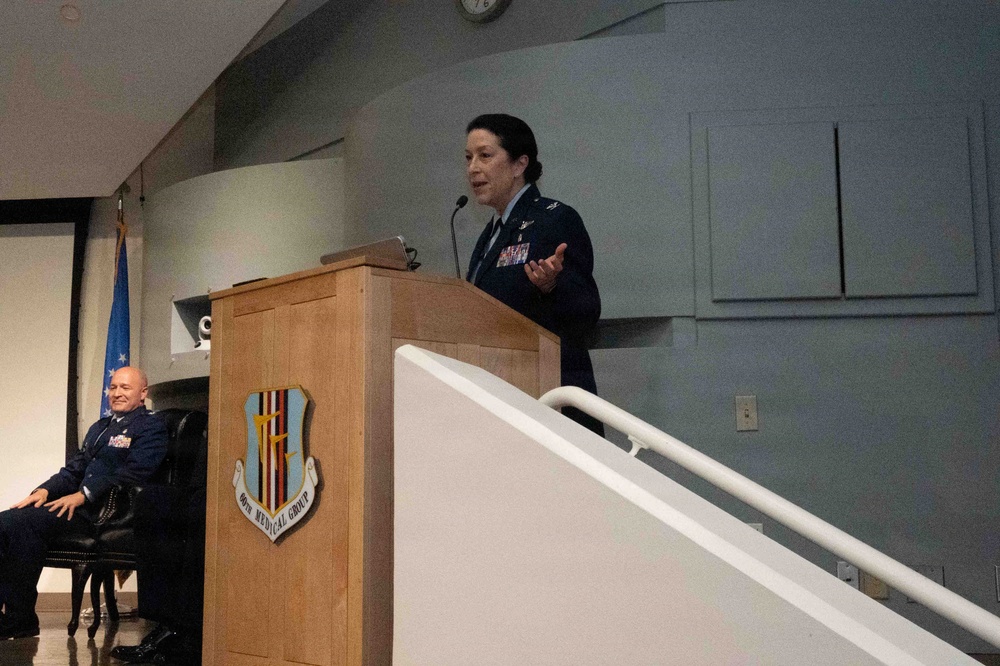 A female military officer in US Air Force dress blues is speaking at a wooden podium bearing the insignia of the 60th Medical Group. A male officer in dress uniform is seated in the background to the left. The setting appears to be an auditorium or formal briefing room.