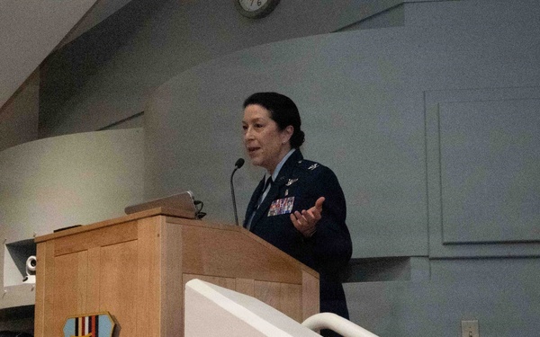 A female military officer in US Air Force dress blues is speaking at a wooden podium bearing the insignia of the 60th Medical Group. A male officer in dress uniform is seated in the background to the left. The setting appears to be an auditorium or formal briefing room.