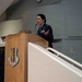 A female military officer in US Air Force dress blues is speaking at a wooden podium bearing the insignia of the 60th Medical Group. A male officer in dress uniform is seated in the background to the left. The setting appears to be an auditorium or formal briefing room.