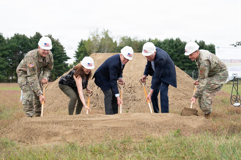 A groundbreaking ceremony is taking place outdoors, with five participants using ceremonial golden shovels to break ground on a new construction project. Two individuals are wearing U.S. Army combat uniforms, while the others are in civilian attire, all wearing white hard hats adorned with American flag stickers. A sign partially visible on the right appears to reference 'Family Readiness,' suggesting a military-affiliated facility.