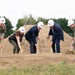 A groundbreaking ceremony is taking place outdoors, with five participants using ceremonial golden shovels to break ground on a new construction project. Two individuals are wearing U.S. Army combat uniforms, while the others are in civilian attire, all wearing white hard hats adorned with American flag stickers. A sign partially visible on the right appears to reference 'Family Readiness,' suggesting a military-affiliated facility.