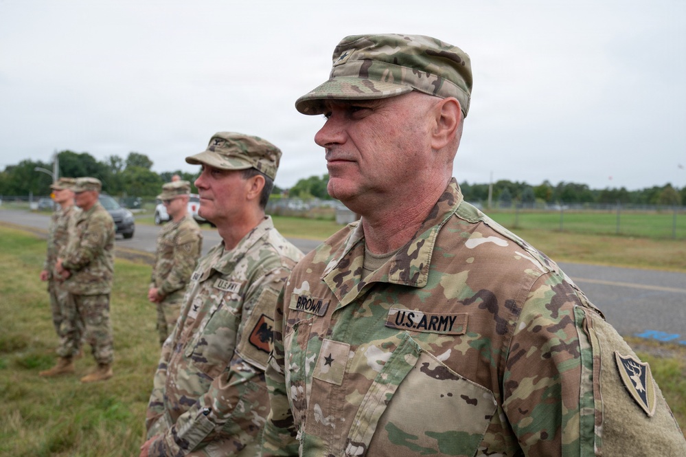 A group of U.S. Army soldiers in Operational Camouflage Pattern (OCP) uniforms are standing in formation outdoors. The foreground soldier's name tape reads 'BROWN' and displays a Brigadier General star insignia on the collar, along with a unit shoulder sleeve insignia. The setting appears to be a military installation with a paved road, fencing, and trees visible in the background.