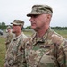 A group of U.S. Army soldiers in Operational Camouflage Pattern (OCP) uniforms are standing in formation outdoors. The foreground soldier's name tape reads 'BROWN' and displays a Brigadier General star insignia on the collar, along with a unit shoulder sleeve insignia. The setting appears to be a military installation with a paved road, fencing, and trees visible in the background.