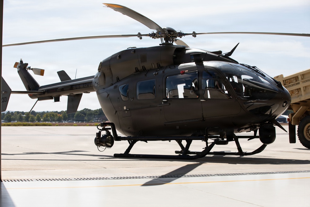A U.S. Army military helicopter, identifiable as a UH-72 Lakota (based on its distinctive shape and 'U.S. ARMY' markings), is parked on a concrete tarmac or flight line. The aircraft is painted in a dark olive/gray military finish and is equipped with an under-nose sensor/camera turret. A military vehicle is partially visible to the right of the frame.