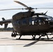 A U.S. Army military helicopter, identifiable as a UH-72 Lakota (based on its distinctive shape and 'U.S. ARMY' markings), is parked on a concrete tarmac or flight line. The aircraft is painted in a dark olive/gray military finish and is equipped with an under-nose sensor/camera turret. A military vehicle is partially visible to the right of the frame.