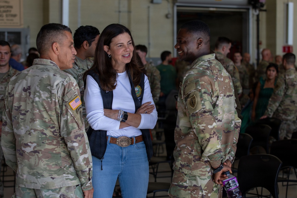 A civilian woman in a white long-sleeve shirt, dark vest, and jeans is engaged in a friendly conversation with two U.S. Army soldiers in OCP (Operational Camouflage Pattern) uniforms. The setting appears to be a military hangar or large indoor facility with numerous other soldiers visible in the background. The interaction appears cordial and informal, with visible smiling among the participants.