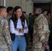 A civilian woman in a white long-sleeve shirt, dark vest, and jeans is engaged in a friendly conversation with two U.S. Army soldiers in OCP (Operational Camouflage Pattern) uniforms. The setting appears to be a military hangar or large indoor facility with numerous other soldiers visible in the background. The interaction appears cordial and informal, with visible smiling among the participants.