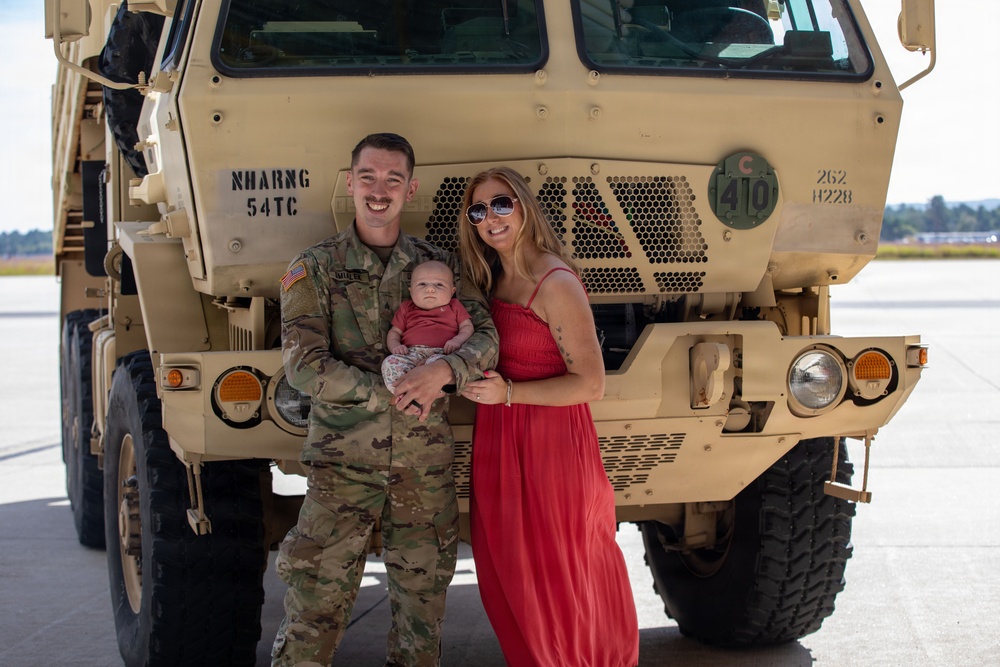 A U.S. Army soldier in OCP (Operational Camouflage Pattern) uniform poses with a woman in a red dress and a newborn infant in front of a tan military tactical truck. The vehicle markings indicate it belongs to the New Hampshire Army National Guard (NHARNG), 54th Transportation Company. The setting appears to be a tarmac or airfield, suggesting a deployment or homecoming event.