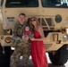 A U.S. Army soldier in OCP (Operational Camouflage Pattern) uniform poses with a woman in a red dress and a newborn infant in front of a tan military tactical truck. The vehicle markings indicate it belongs to the New Hampshire Army National Guard (NHARNG), 54th Transportation Company. The setting appears to be a tarmac or airfield, suggesting a deployment or homecoming event.