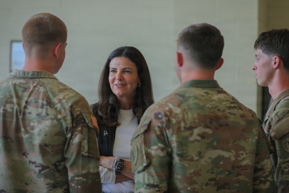 A civilian woman is engaged in conversation with three military personnel wearing OCP (Operational Camouflage Pattern) uniforms. The setting appears to be an indoor military facility or building. The interaction appears cordial and professional in nature.
