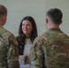 A civilian woman is engaged in conversation with three military personnel wearing OCP (Operational Camouflage Pattern) uniforms. The setting appears to be an indoor military facility or building. The interaction appears cordial and professional in nature.