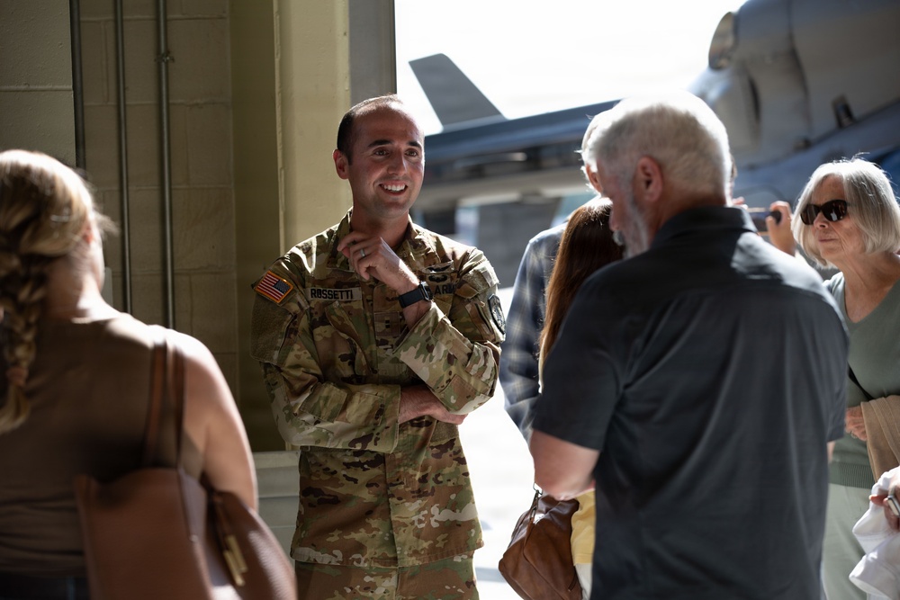 A U.S. Army soldier named Rossetti, wearing OCP (Operational Camouflage Pattern) uniform, is smiling and conversing with a group of civilians inside or near a military hangar. Military aircraft are visible in the background through the hangar opening. The atmosphere appears relaxed and social, suggesting a family day, open house, or homecoming event.