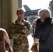 A U.S. Army soldier named Rossetti, wearing OCP (Operational Camouflage Pattern) uniform, is smiling and conversing with a group of civilians inside or near a military hangar. Military aircraft are visible in the background through the hangar opening. The atmosphere appears relaxed and social, suggesting a family day, open house, or homecoming event.