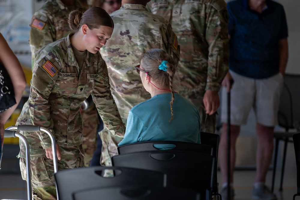 A U.S. Army soldier, identified by name tape as 'CRESSWELL' and wearing OCP (Operational Camouflage Pattern) uniform, is leaning down to speak with an elderly woman seated in a wheelchair. The interaction appears to be a welfare or assistance engagement, likely during a humanitarian or disaster relief operation. Several other soldiers in OCP uniforms are visible in the background.