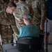 A U.S. Army soldier, identified by name tape as 'CRESSWELL' and wearing OCP (Operational Camouflage Pattern) uniform, is leaning down to speak with an elderly woman seated in a wheelchair. The interaction appears to be a welfare or assistance engagement, likely during a humanitarian or disaster relief operation. Several other soldiers in OCP uniforms are visible in the background.