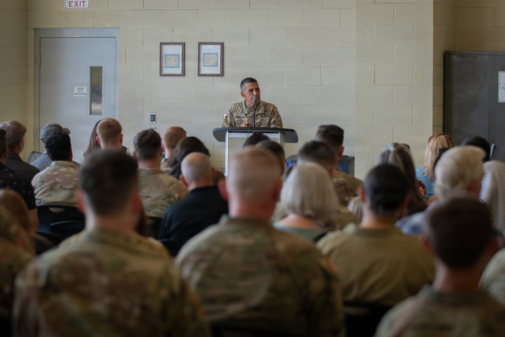 A military officer in camouflage uniform stands at a podium addressing a large seated audience in what appears to be a military facility or armory. The audience consists of a mix of uniformed military personnel and civilians seated in rows facing the speaker. The setting suggests a formal briefing, ceremony, or unit assembly.