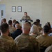 A military officer in camouflage uniform stands at a podium addressing a large seated audience in what appears to be a military facility or armory. The audience consists of a mix of uniformed military personnel and civilians seated in rows facing the speaker. The setting suggests a formal briefing, ceremony, or unit assembly.