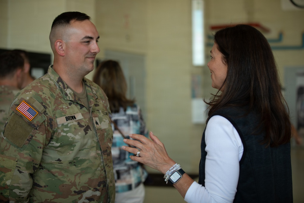 A U.S. Army soldier in OCP (Operational Camouflage Pattern) uniform with the name tape 'BROWN' is engaged in conversation with a civilian woman wearing a white long-sleeve shirt and dark vest. The interaction appears cordial and informal, taking place inside what appears to be a community or military facility. Other individuals are visible in the blurred background.