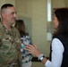 A U.S. Army soldier in OCP (Operational Camouflage Pattern) uniform with the name tape 'BROWN' is engaged in conversation with a civilian woman wearing a white long-sleeve shirt and dark vest. The interaction appears cordial and informal, taking place inside what appears to be a community or military facility. Other individuals are visible in the blurred background.