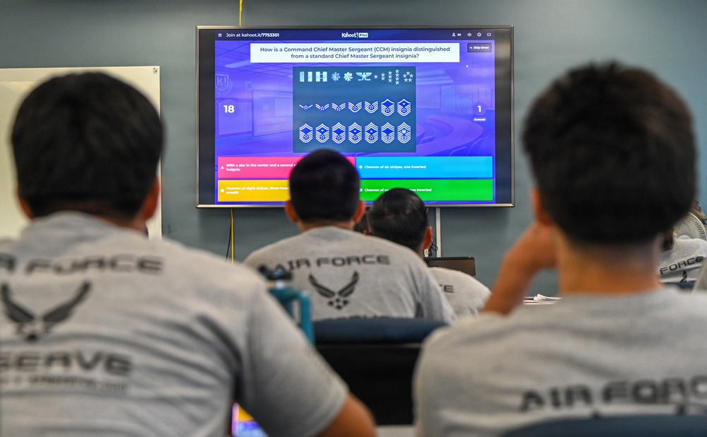 A group of U.S. Air Force Reserve personnel are participating in a Kahoot! quiz session focused on military insignia knowledge. The screen displays a question about distinguishing a Command Chief Master Sergeant (CCM) insignia from a standard Chief Master Sergeant insignia. Participants are seated at desks wearing matching gray Air Force Reserve t-shirts and facing the display screen.