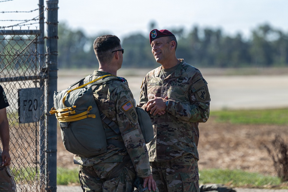 Two U.S. Army soldiers in MultiCam/OCP camouflage uniforms are engaged in conversation near a chain-link fence with barbed wire at what appears to be an airfield or military installation. The soldier on the right is wearing a maroon/crimson beret, indicative of airborne-qualified personnel, while the soldier on the left is carrying a military parachute pack with yellow static line. The setting suggests a pre- or post-jump airborne operation.