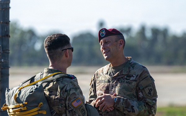 Two U.S. Army soldiers in MultiCam/OCP camouflage uniforms are engaged in conversation near a chain-link fence with barbed wire at what appears to be an airfield or military installation. The soldier on the right is wearing a maroon/crimson beret, indicative of airborne-qualified personnel, while the soldier on the left is carrying a military parachute pack with yellow static line. The setting suggests a pre- or post-jump airborne operation.