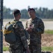 Two U.S. Army soldiers in MultiCam/OCP camouflage uniforms are engaged in conversation near a chain-link fence with barbed wire at what appears to be an airfield or military installation. The soldier on the right is wearing a maroon/crimson beret, indicative of airborne-qualified personnel, while the soldier on the left is carrying a military parachute pack with yellow static line. The setting suggests a pre- or post-jump airborne operation.