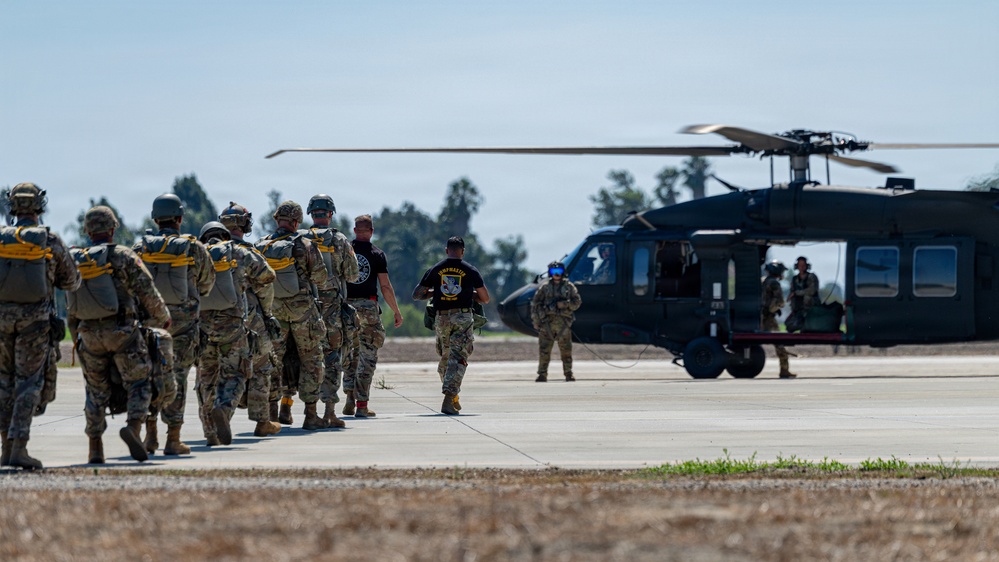 A group of military personnel in full combat gear and parachute equipment are lined up on a tarmac, preparing to board a dark military helicopter, likely a UH-60 Black Hawk. Two individuals in black t-shirts appear to be instructors or jumpmaster personnel guiding the formation toward the aircraft. The scene depicts a pre-jump or air assault operation at a military airfield.
