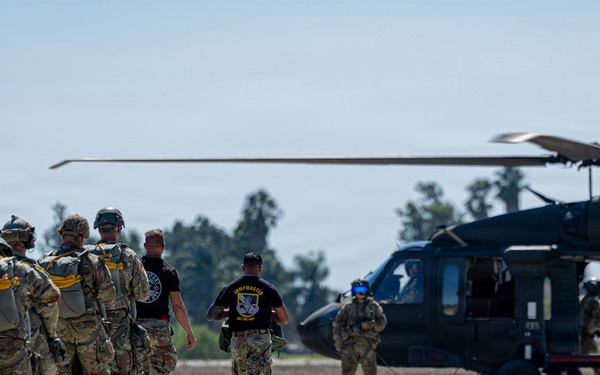A group of military personnel in full combat gear and parachute equipment are lined up on a tarmac, preparing to board a dark military helicopter, likely a UH-60 Black Hawk. Two individuals in black t-shirts appear to be instructors or jumpmaster personnel guiding the formation toward the aircraft. The scene depicts a pre-jump or air assault operation at a military airfield.